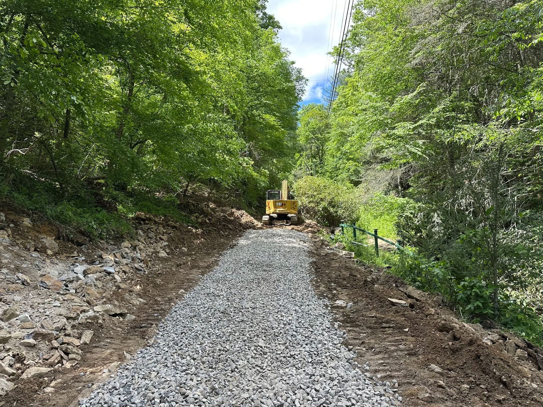 A freshly graveled access road cut through the woods, with an excavator still working at the far end.