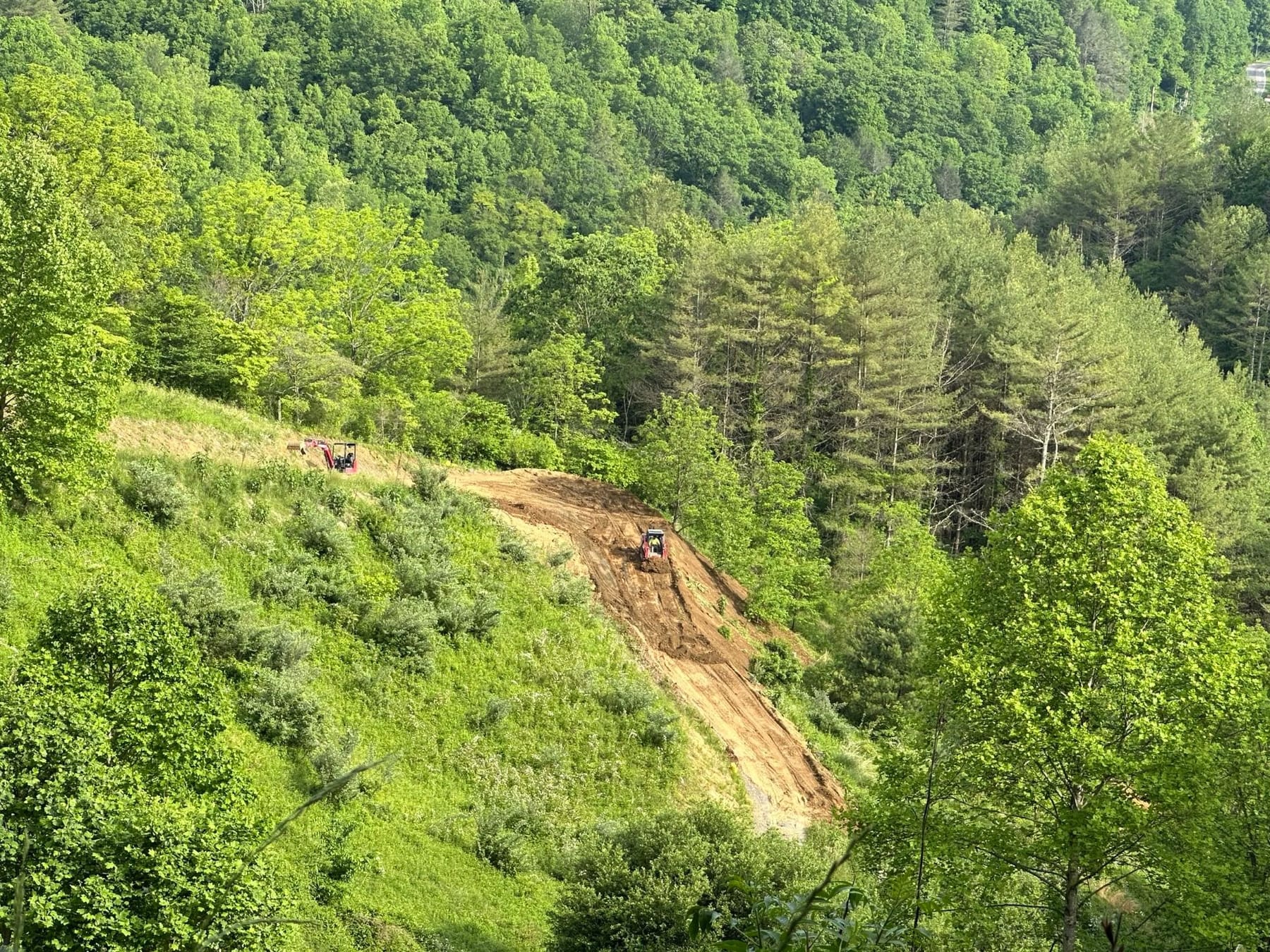 A new cut graded into a steep, wooded mountainside, with a track loader on the slope and a mini excavator up top.