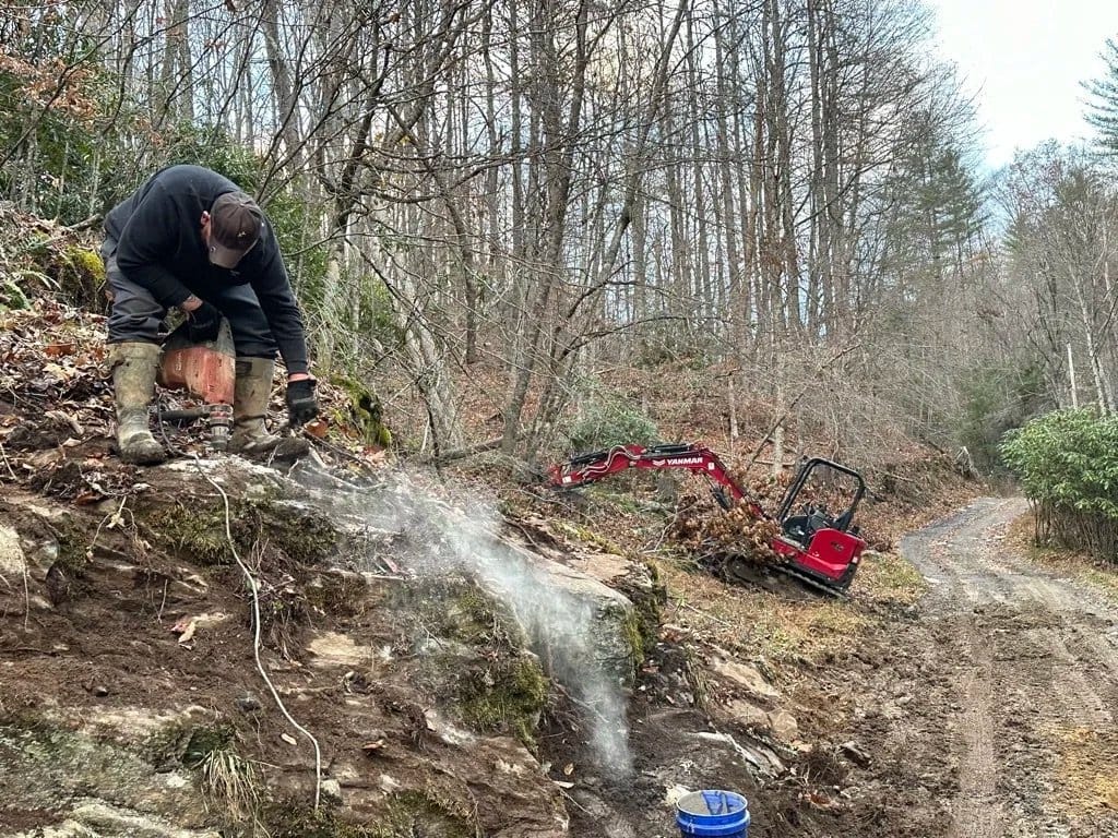 Nute Nicholson splitting rock on a hillside above an access road, with a mini excavator standing by.