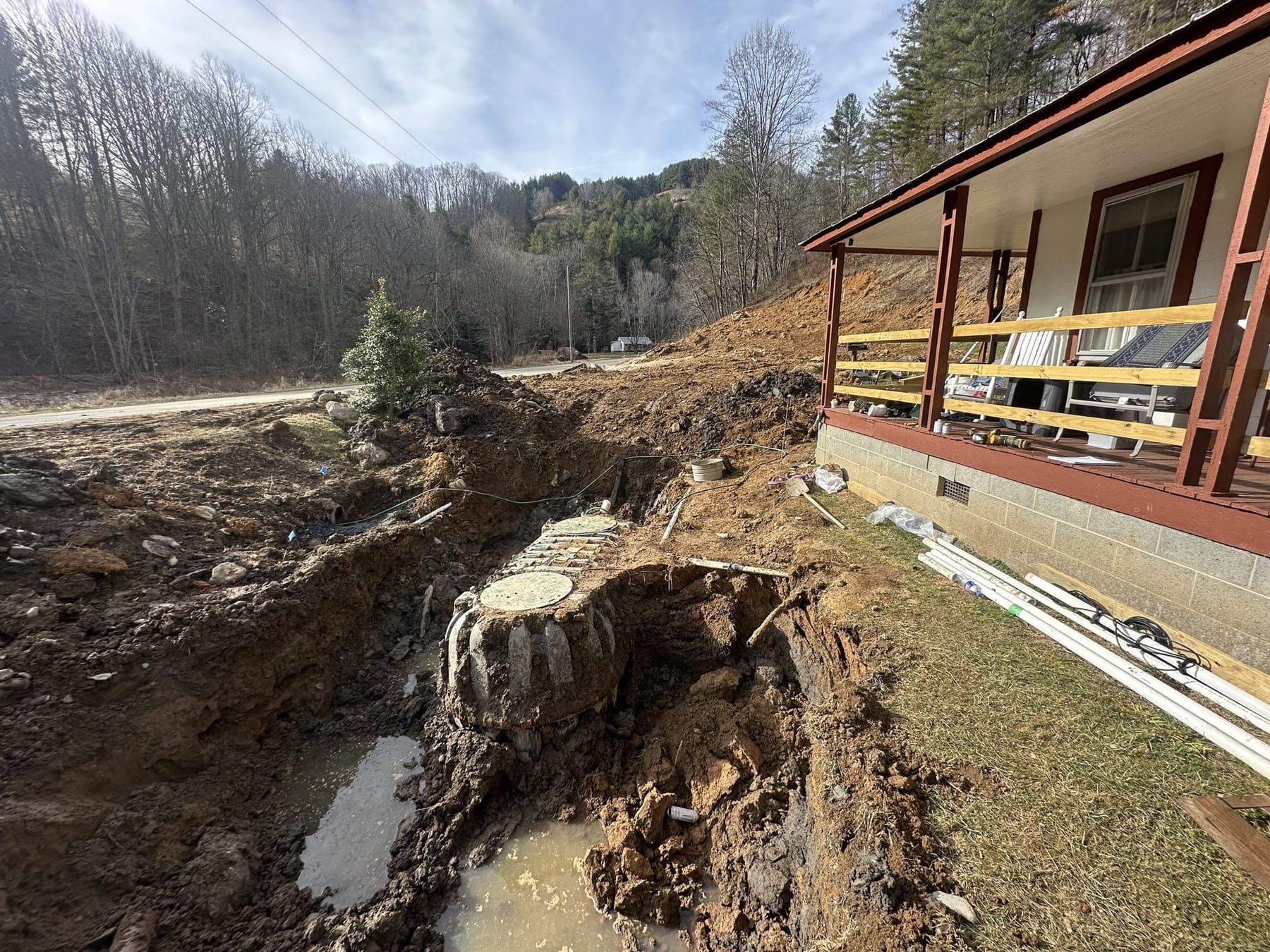 An excavation opened up beside a mountain home, with water in the trench and the septic tank exposed.