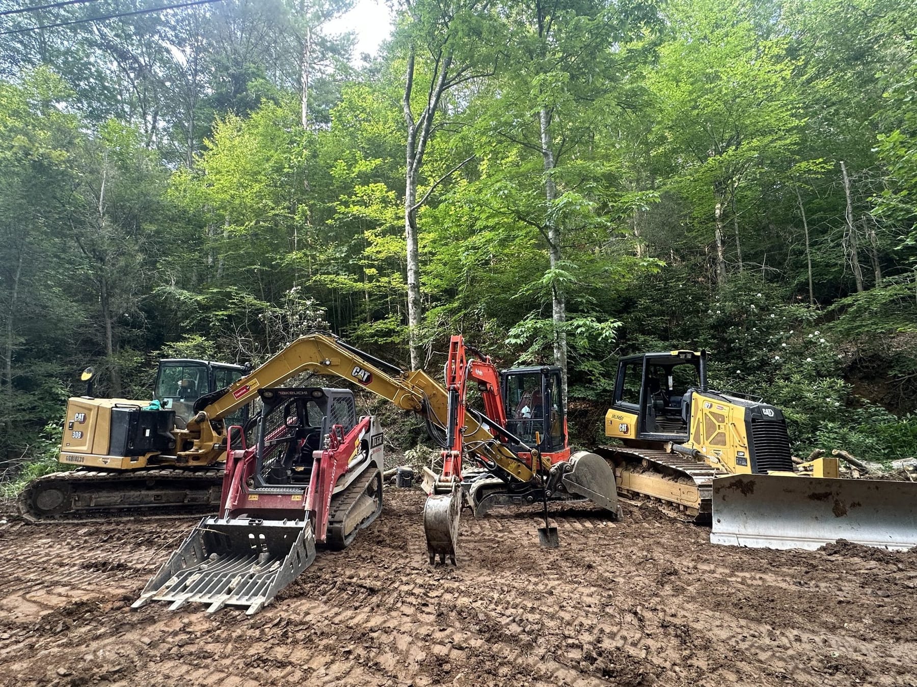 A CAT excavator, a Takeuchi track loader, a red mini excavator and a CAT dozer lined up on a wooded mountain site.