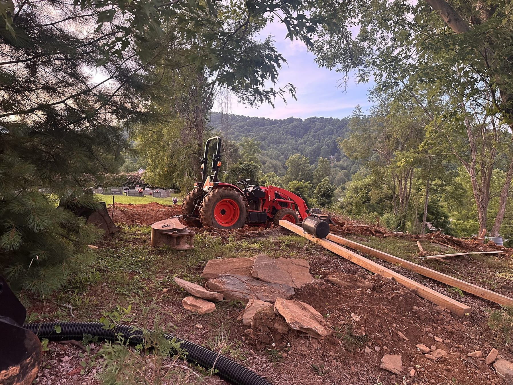 A tractor at work on a newly cleared mountain lot, with drain pipe and timbers staged on the opened ground.
